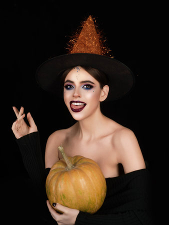 Witch posing with pumpkin in black studio. Young woman in witch hat celebrating Halloween. Sexy...