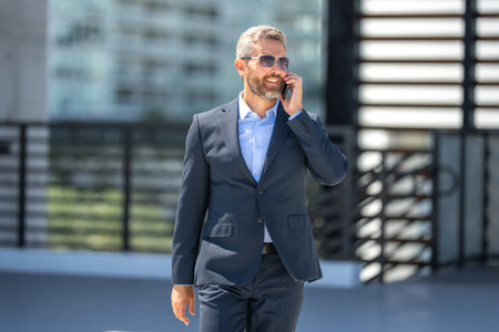 Businessman walking and talking on phone. Confident business man outdoors with phone. Business phone conversation. Busy corporate leader talking on phone. Rich business man in suit. Business call.の写真素材
