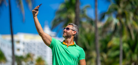 Tourist takes a selfie near palm tree. Man captures a selfie during summer vacation. Traveler snaps a selfie in a tropical destination. A selfie with palm trees captures the tropical vibes.の写真素材
