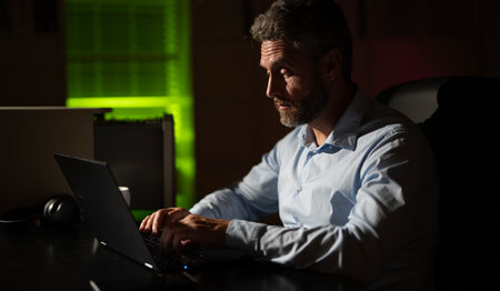 Tired businessman working late at night on a laptop in a dark office. Bored manager struggling with...