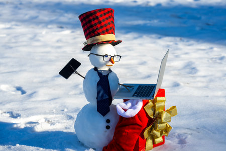 Snowman with mobile phone and laptop during winter celebration. Snowman working online in winter. Online greeting for New Year. Holiday snowman connecting to internet in snowfield.の写真素材