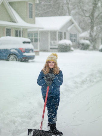 Funny Kid removing snow with a shovel in the yard on snowy winter day. Child clearing sidewalks and shoveling snow. Child removing snow with shovel. Little helper. Winter kids fun outdoor.の写真素材
