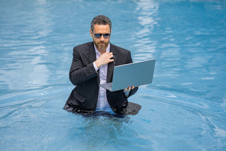 Funny corporate leader swimming. Remote job by the beach. Luxury vacation with laptop. Freelance work-life. Business humor. Handsome employee in suit in water. Relaxing after job. Success in career.の写真素材