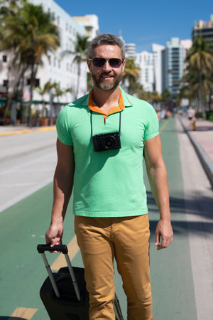 A traveler with camera and luggage on streets of Miami. A man traveler enjoys his summer vacation in a tropical city. A traveler tourist with suitcase on summer city. Man traveler walk on city.の写真素材