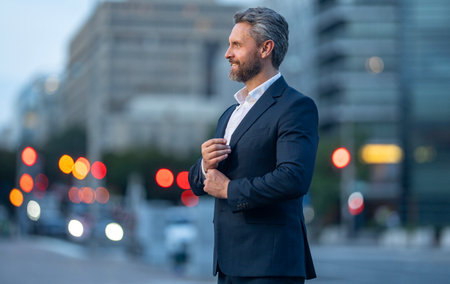 Happy mature businessman standing outside in an urban city street. Man in suit outdoor city night portrait. Mature business man walking on the city. Office worker posing on a city street.の写真素材