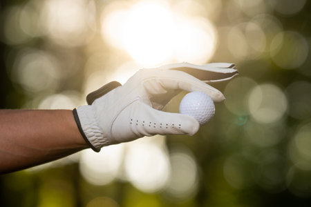 Golfer hand holds a ball closeup on a green background. Golf ball background concept. Closeup of a hand with a golf ball on a sunny summer course. Golfing and recreation. Golf concept.の写真素材