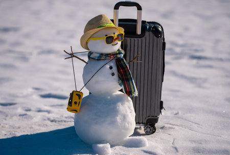 Snowman with suitcase ready for holiday trip. Winter vacation starts with snowman. Christmas snowman holding travel bag outdoors. Festive snowman tourist on snowy adventure. New Year journey.の写真素材