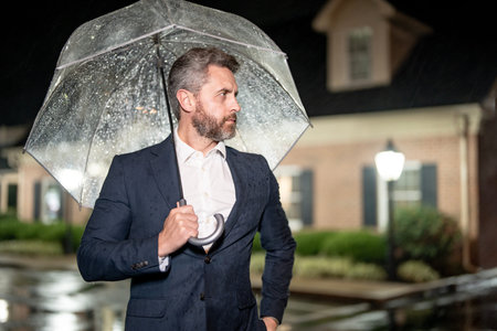 Businessman with umbrella in rain night. Business patience. Businessman waiting in rain. Businessman on night street. Man in suit patience while waiting near office. Man waiting in rainy weather.の写真素材