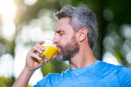 A healthy man holds a glass of fresh juice outdoors. Man drink orange juice as a natural supplement after a workout. Sport man drink glass of juice. A man holds a fresh juice as part of healthy diet.の写真素材