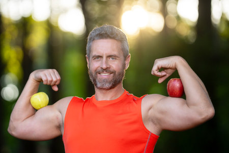 A strong man flexes his biceps with apples. Mature athlete shows biceps during a workout in the park. A middle-aged sportsman trains biceps. Male biceps after exercise.の写真素材