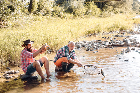 Two men friends fishing. Flyfishing angler makes cast, standing in river water. Old and young fisherman.の写真素材