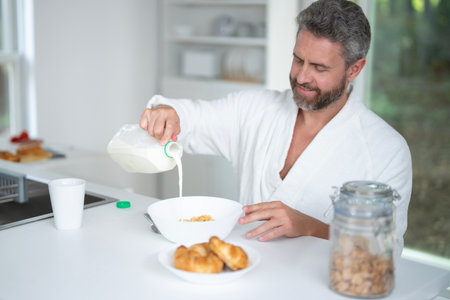 Man eats breakfast in the kitchen. Guy enjoys fresh food at the kitchen table in the morning. A man eats cereal and fruit in the kitchen. Man eating healthy food in the kitchen.の写真素材