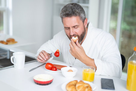 Man eats breakfast in the kitchen on morning. Man 40s enjoys a healthy meal at the kitchen table. A man eats cereal and fruit in the kitchen during morning hours. Man eats vegetables for lunch.の写真素材