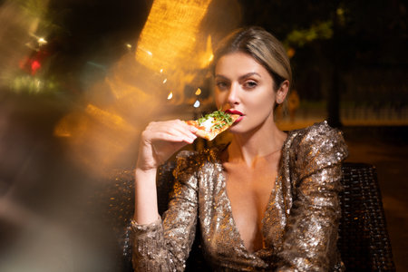Elegant woman eating pizza in night cafe. Sensual woman enjoying food in luxury setting. Woman posing with gourmet pizza slice. Sensual expression in city restaurant.の写真素材