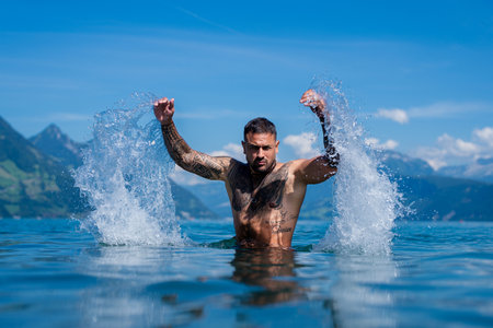 Fit tattooed man trains in water. Summer swim. Splash effect around body. Man splashes water with power. Tattooed athlete man enjoys summer swim outdoors. Strong sexy male body.の写真素材