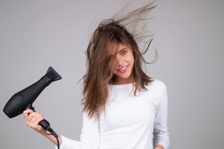A woman dries her hair with a blow dryer and smiles. Hair routine with brush and dryer. Beauty model hold hair dryer. Haircare concept.の写真素材