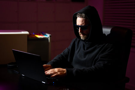 Hacker with laptop in a dark office. Anonymous hacker in a hoodie hacking data late at night. Cyber criminal and hacker stolen digital information concept. Hacker using laptop in night office.の写真素材