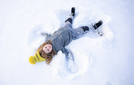 Happy child making a snow angel in winter. Kid playing in snow outdoors. Child lying in snow making angel wings shape. Angel kid play in winter. Winter fun with a happy child making snow angels.の写真素材