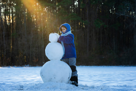 Kid building a snowman on winter day. Childhood and winter fun. Kid creating snowman outdoors. Child enjoying snow winter fun and making snowman. Child playing with snowman.の写真素材