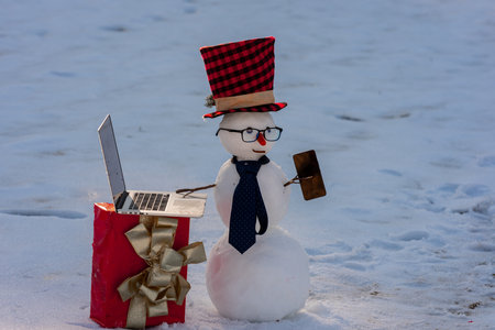 Snowman talking on smartphone in snowy landscape. Snowman making a business phone call. Funny snowman using laptop outdoors. Snowman with mobile phone during winter holiday.の写真素材