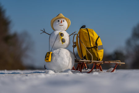 Snowman traveler with suitcase outdoor. Winter tour with snowman and backpack. Snowman posing with travel bag in cold weather. Snow man enjoying vacation in the snow. Snowman with sleigh and luggage.の写真素材