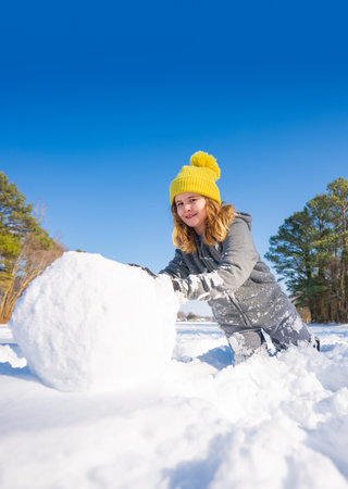 Happy kid rolling a big snowball on a sunny winter day. Smiling kid playing in snow outdoors. Cute...