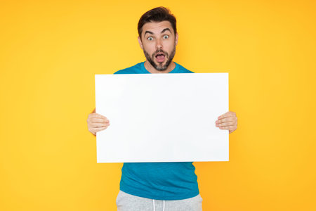 Amazed handsome man with blank banner ad isolated on studio background. Portrait of surprised man holding empty blank poster. Amazed man showing blank poster signboard, billboard or banner.の写真素材