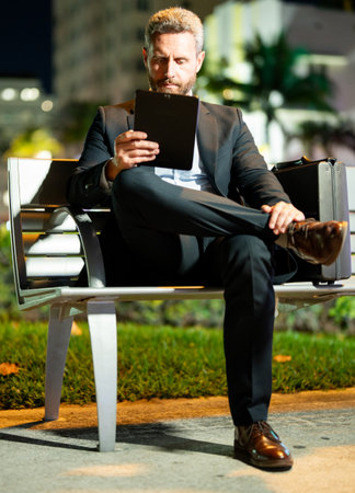 Outdoor business. Serious business man working on tablet outdoors. Businessman with tablet outdoor. Businessman with a tablet on night street. Businessman using tablet, sit on bench. Night business.の写真素材