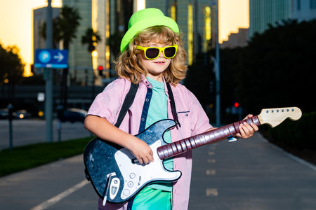 Young kid performing music in city. Little musician in sunglasses play electric guitar. Child rocking with guitar. Kid enjoying street music performance. Kid with electric guitar. Fun young guitarist.の写真素材