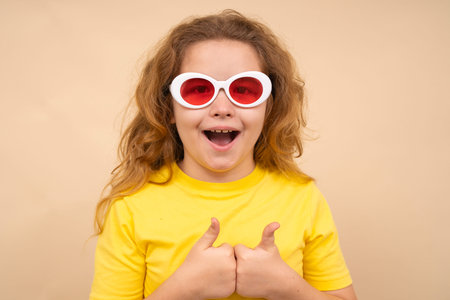 Kid with sunglasses shows thumbs-up. Child with sunglasses smiles brightly isolated on studio...