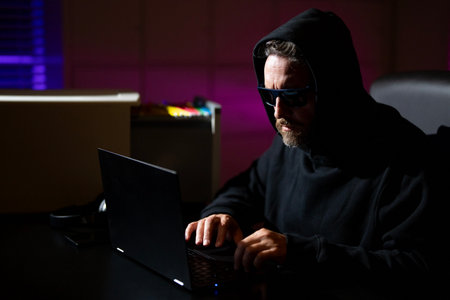 Hacker types on a laptop in a night office during a cyber attack. Hacker and stolen financial data concept. Digital money hacker. Cyber attack. Hacker in a hoodie using laptop in office.の写真素材