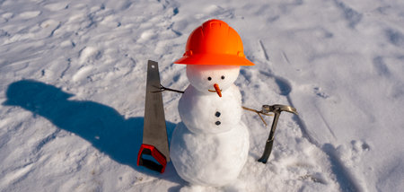 Snowman builder with construction equipment tools in snow. Snow man worker hold saw and hammer. Winter construction concept. Winter construction site greeting card. Snowman construction worker.の写真素材