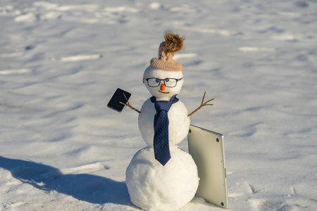Businessman snowman talking on smartphone in snow. Snowman making a business phone call. Funny snowman hacker using laptop outdoors. Snowman with mobile phone during winter holiday.の写真素材