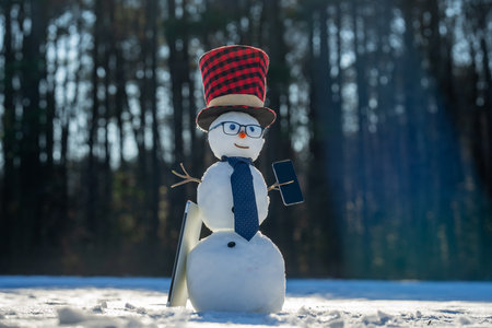 Snowman making a phone call with smartphone. Snow man using laptop outdoors in snow. Funny snowman business man with mobile phone. Winter celebration with snowman.の写真素材