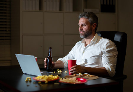 Businessman working late at office desk. Employee after night work. Overtime at night office. Office worker finishing work on laptop after hours. Overworked man eating during night work.の写真素材
