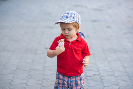 A cute baby enjoys eating ice cream outdoors. Child holds a cone on a street. Little kid tastes ice cream. Happy childhood. Summer kids snack. Baby smiles with ice cream.の写真素材