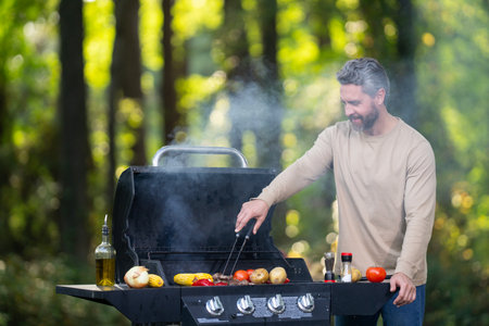 Handsome man grilling meat on a backyard grill. Fit male cooking steak on an outdoor grill. Mature man barbecuing food. Man with grill on nature. BBQ grilling outdoor. Backyard grill.の写真素材