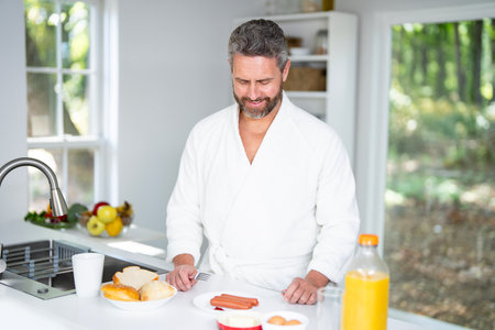 Man eats breakfast in the kitchen on a morning. A man enjoys fresh food at the kitchen table. A man eats cereal and fruit in the kitchen. Guy enjoys eggs and vegetables at the modern kitchen.の写真素材