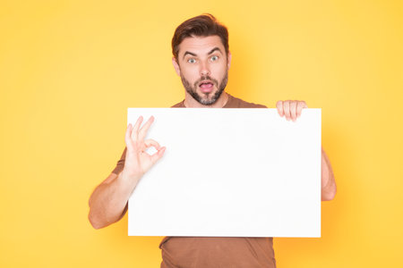 Guy with blank banner ad, posing isolated over studio background. Portrait of attractive man holding empty blank poster. Man showing ok sign and blank poster, gesturing ok finger. Billboard or banner.の写真素材
