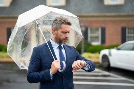 Businessman waits under the rain holding an umbrella. A confident man checks his watch while...