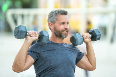 Sporty man lifting dumbbells outdoors. Sport man exercising with dumbbells in public outdoor gym. Fit guy working out in the park. Sportsman lifting dumbbells. Middle aged man training with dumbbells.の写真素材