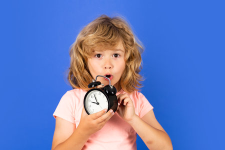 Blonde child holding alarm clock with funny face. Morning school time concept with surprised kid. Portrait of kid with big clock and shocked expression. Alarm ringing while kid looks confused.の写真素材