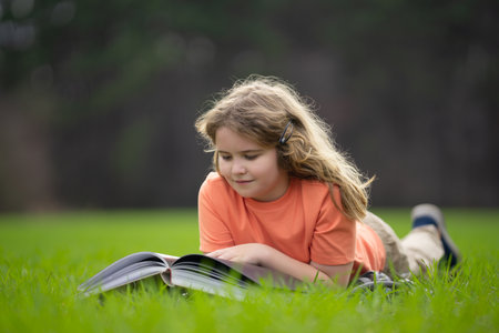 Kid read a book in the grass. Kid read a book story outdoors. Kid read book in the meadow on a sunny day. Reading in nature. Schoolboy with book. Kid outdoor learning. Kids read story.の写真素材
