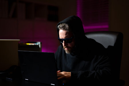 hacker types on a laptop in a dark office. Anonymous hacker in a hoodie hacking data late at night....