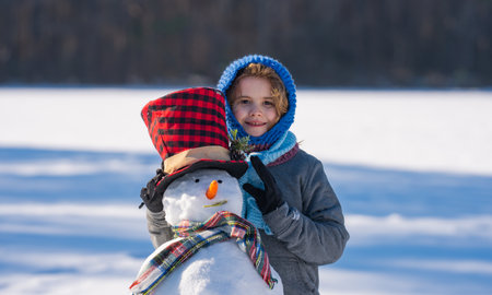 Child hug snowman. Child play with snowman in snow field. Kids winter activity. Child enjoying winter creativity in snowy field. Child decorating snowman. Kid building snowman. Kids winter fun.の写真素材