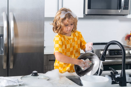 Child washing pan with soap foam. Kid play with soap bubbles at kitchen. Kid washing dishes. Child fun with dish soap at sink.の写真素材
