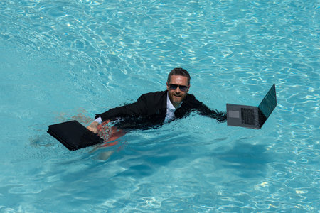 Funny corporate leader swimming. Remote job by the beach. Luxury vacation with laptop. Freelance work-life. Business humor. Handsome employee in suit in water. Relaxing after job. Success in career.の写真素材