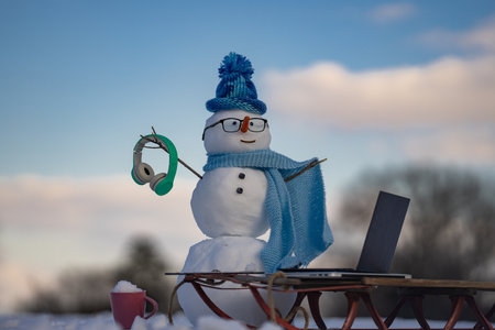 Snowman with headphones and laptop in snow. Winter online shopping. Creative winter snowman with computer setup. Snowman doing remote work in winter landscape. Winter snowman with laptop and sled.の写真素材
