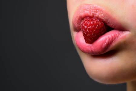 Close up of tongue holding a fresh raspberry. Creative macro concept with berry in mouth. Beauty on lip. Plump lips close up. A raspberry the tongue. Sensual bite. Long tongue.の写真素材