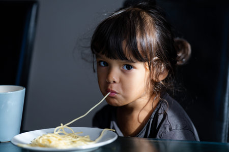 Cute girl slurping noodles in kitchen. Child eating spaghetti with hungry expression. Healthy meal with child. Kid eat spaghetti. Toddler eating pasta lunch indoors. Little child girl tasting pasta.の写真素材